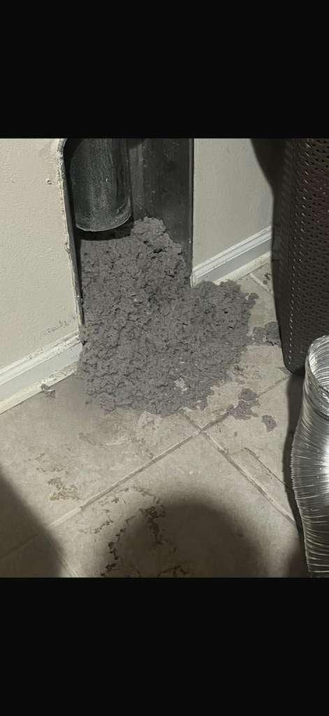 A dense pile of grey dryer-vent lint pulled from a vent opening onto the floor beside a laundry basket