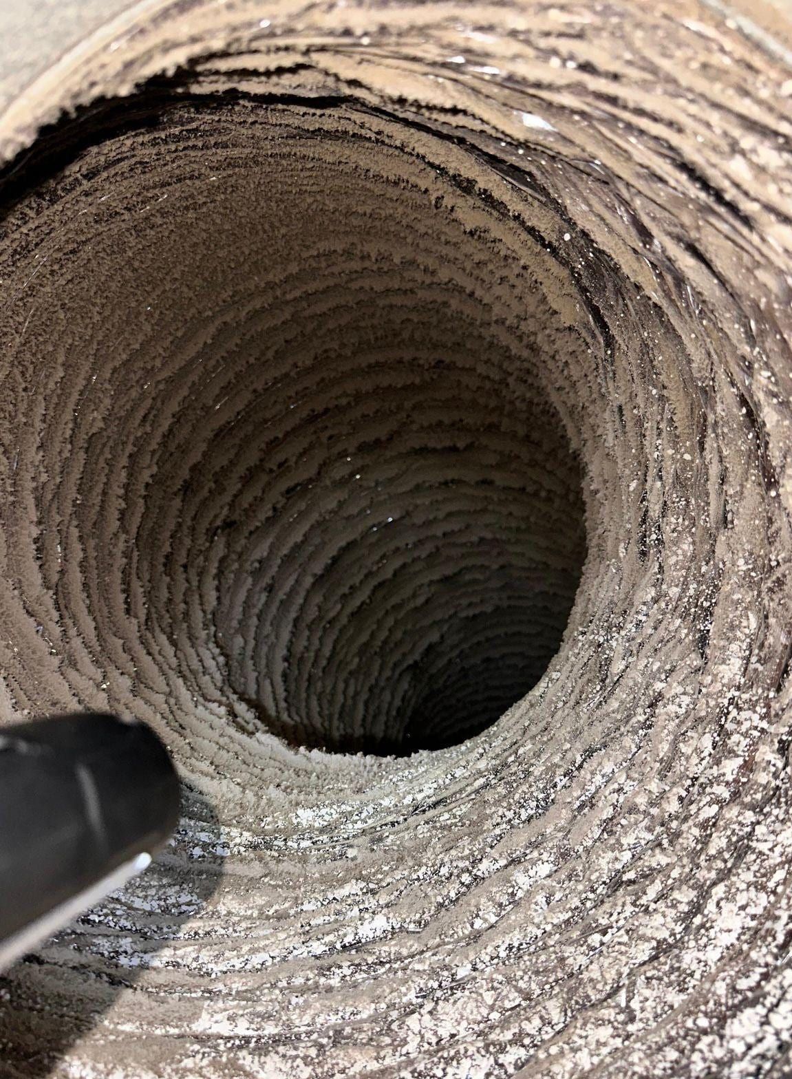 Flexible duct interior coated in thick grey dust, with the dark tip of a cleaning tool entering from the lower left