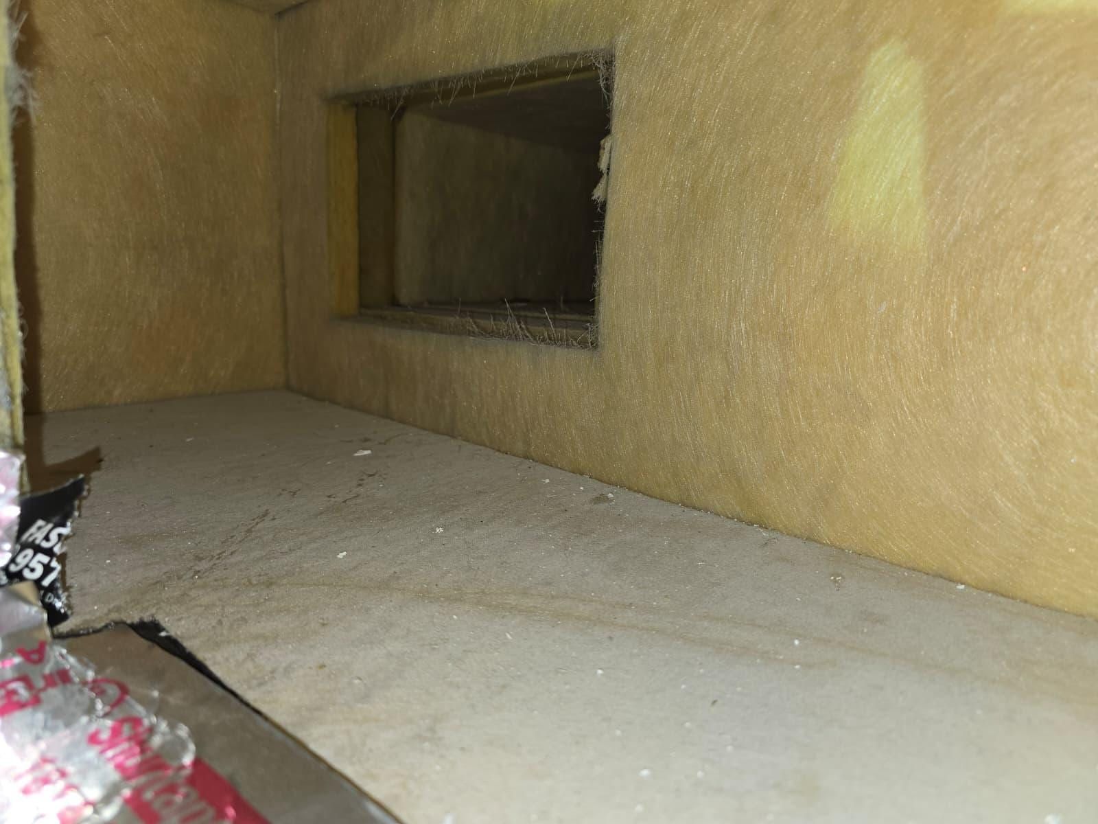 The inside of a fibreglass-lined plenum box, a rectangular duct opening cut into its wall and dust lying on the floor of the box