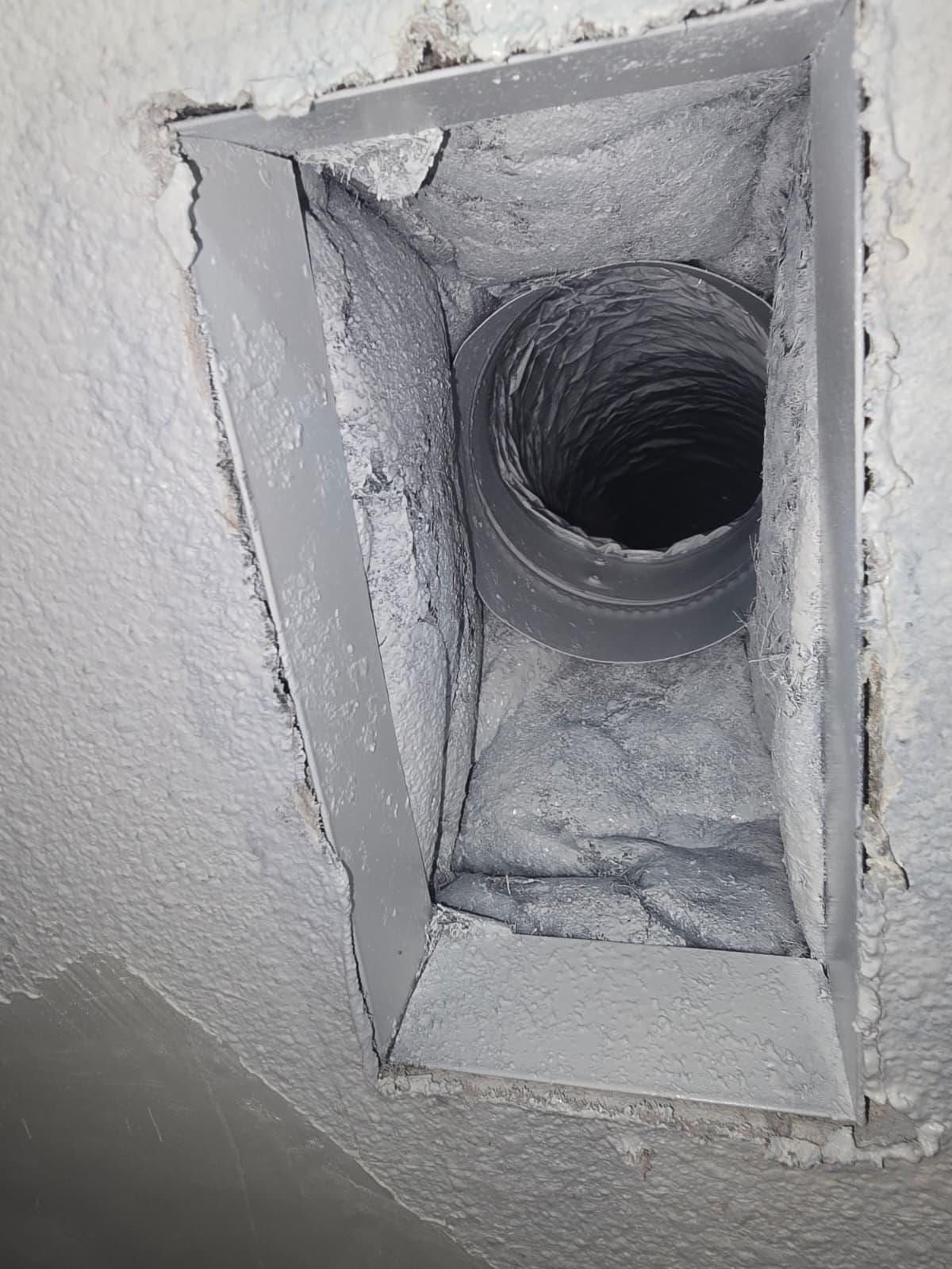 A grey ceiling supply boot opened in a white textured ceiling, its round metal collar and flexible duct furred with grey dust and lint