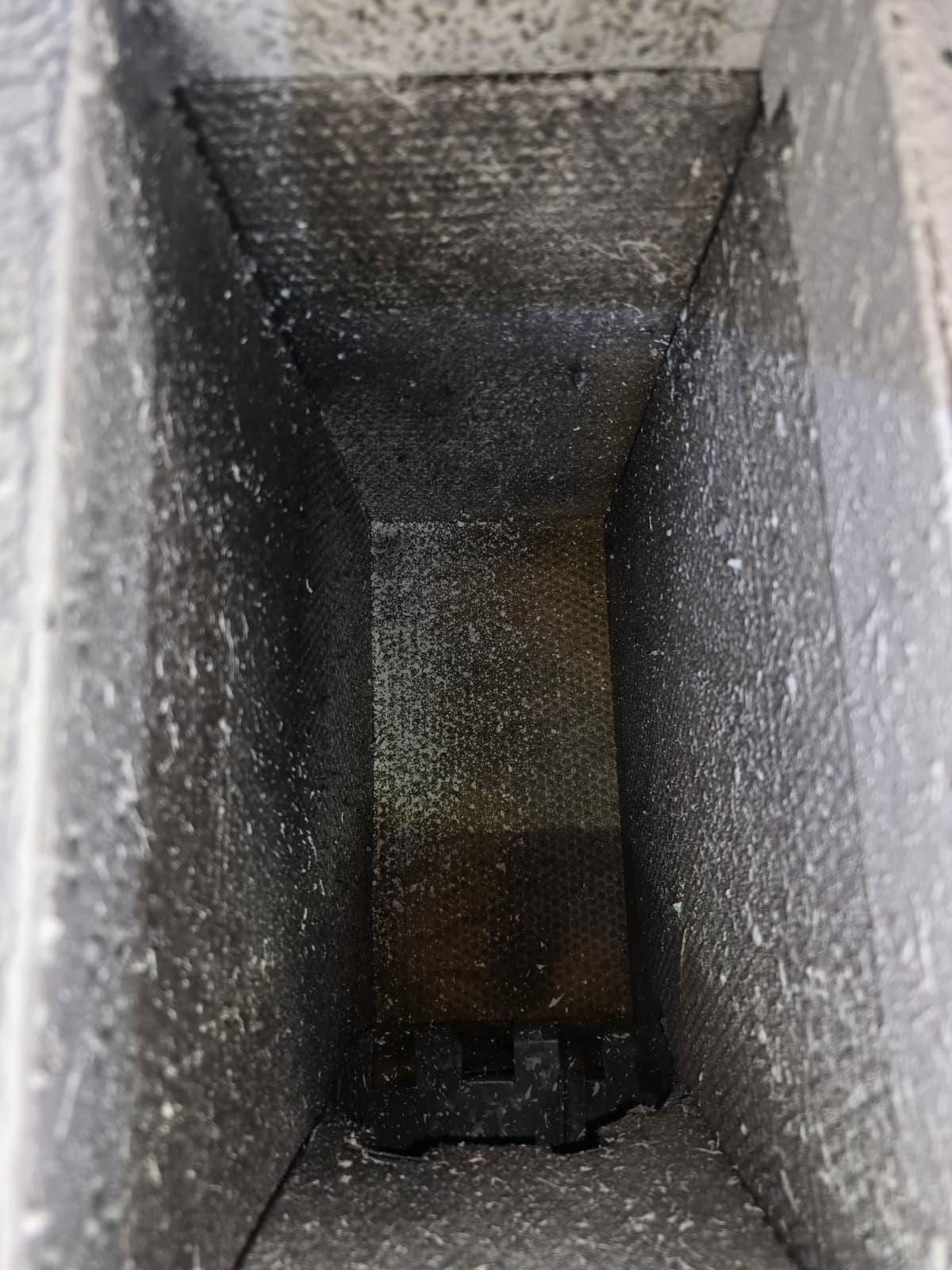 A narrow rectangular duct interior heavily coated in textured grey dust, tapering to a dark gasketed opening