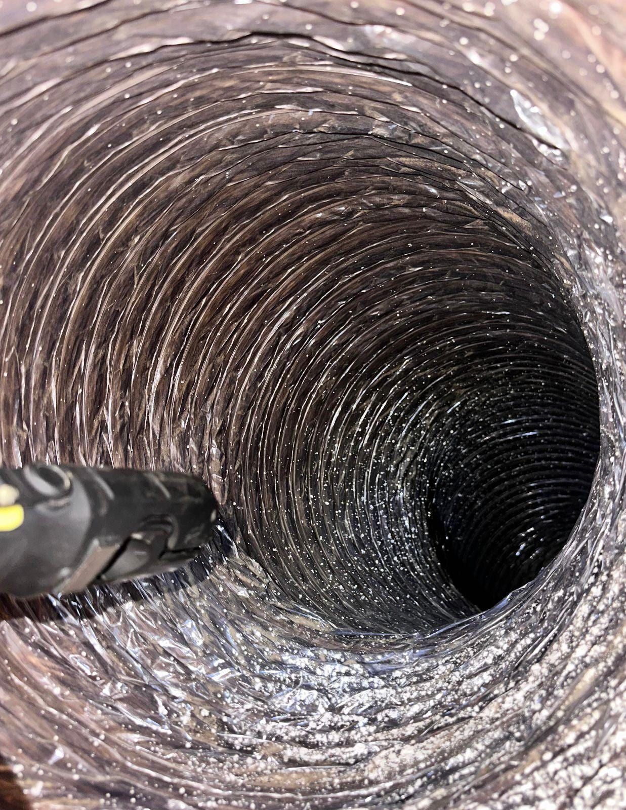 The black head of a cleaning tool entering a dust-coated flexible duct, its ribbed walls spiralling away into darkness