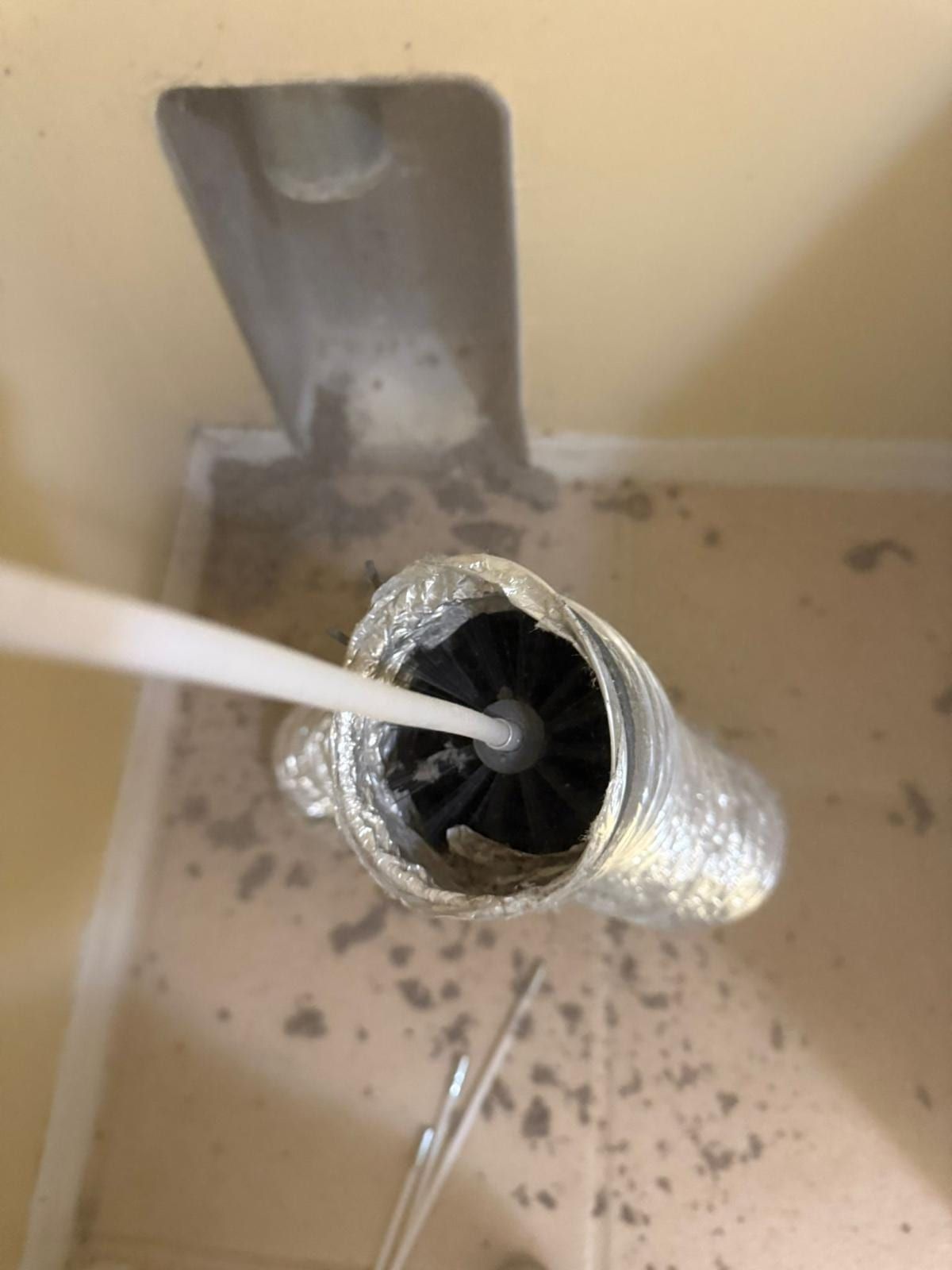 A brush tool and vacuum hose worked into an open flexible duct run, dust visible on the surrounding board and floor